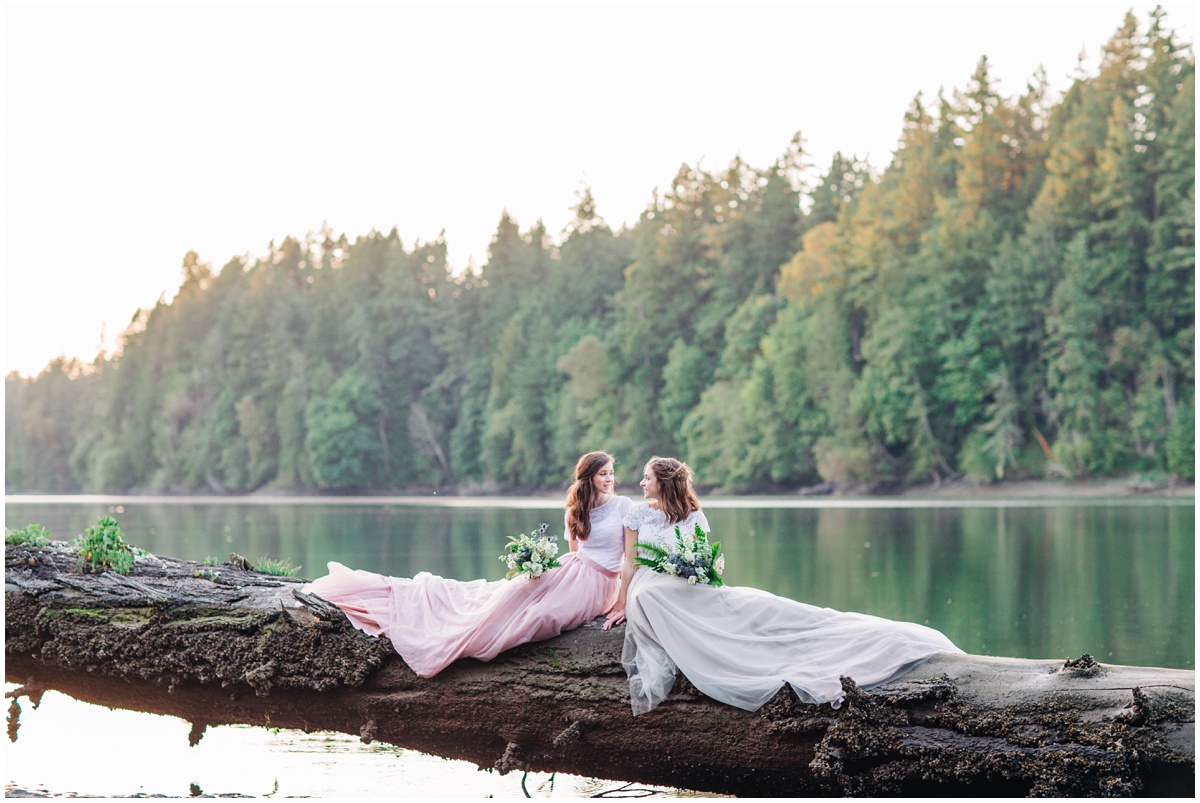 two brides sit on beach log with long gowns